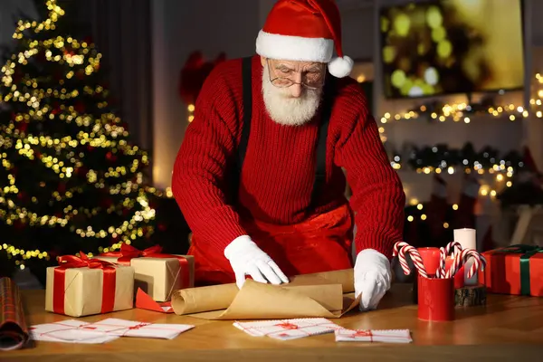 Santa Claus wrapping gift at his workplace in room decorated for Christmas