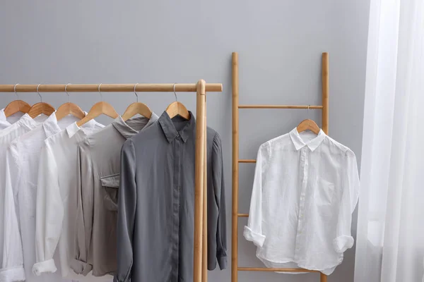 Different stylish shirts hanging on ladder and rack near grey wall in room. Organizing clothes