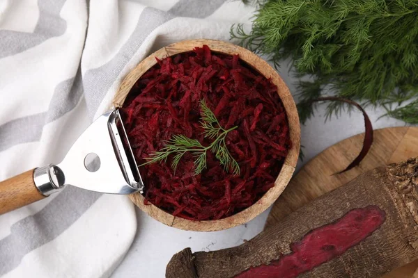 Grated red beet with dill in bowl and vegetable peeler on table, flat lay