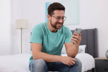 Mutlu adam yatak odasında akıllı telefondan görüntülü konuşma yapıyor.