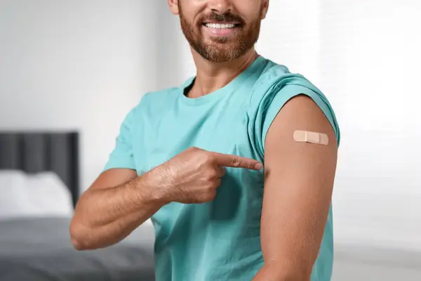 Man pointing at sticking plaster after vaccination on his arm in bedroom, closeup