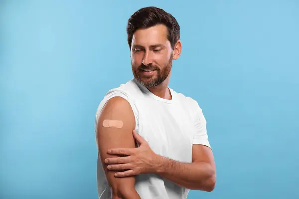 Man with sticking plaster on arm after vaccination against light blue background