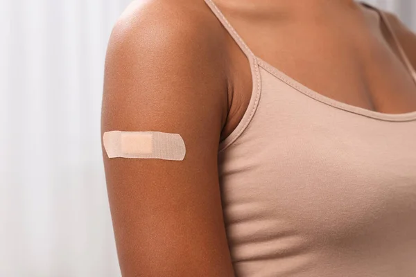 Young woman with adhesive bandage on her arm after vaccination against blurred background, closeup