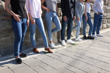 Güneşli bir günde, taştan duvarın yanında şık kotlar giyen bir grup insan yakın çekim yapıyor.