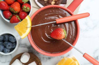 Erimiş çikolatalı fondü kabı, taze portakal, beyaz mermer masa üzerinde farklı meyveler ve çatallar, düz uzanmış.