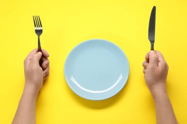 Çatallı, bıçaklı ve boş tabaklı adam sarı masada, üst tarafta.