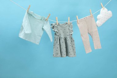 Farklı bebek kıyafetleri ve bulut şekilli yastık açık mavi arka planda çamaşır ipinde kuruyor.