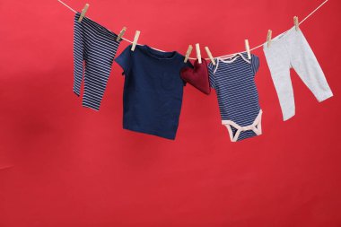 Farklı bebek kıyafetleri ve kalp şeklinde yastıklar kırmızı arka planda çamaşır ipinde kuruyor.
