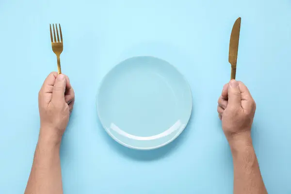 Man with cutlery and empty plate at light blue table, top view