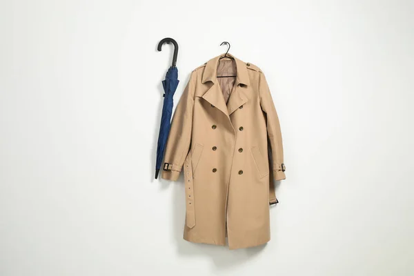 Hanger with beige trench coat and blue umbrella on white wall, space for text