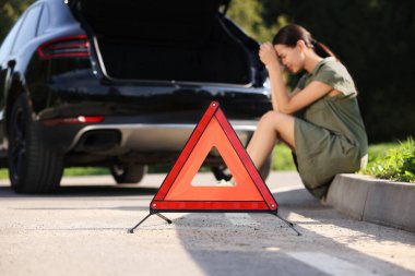 Yol kenarında kırık bir arabanın yanında oturan kadın uyarı üçgenine odaklanıyor.
