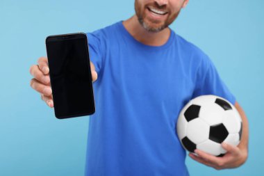 Futbol topu ve açık mavi arka planda akıllı telefonu olan mutlu bir taraftar, yakın plan.
