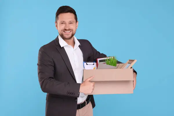 Happy unemployed man with box of personal office belongings on light blue background