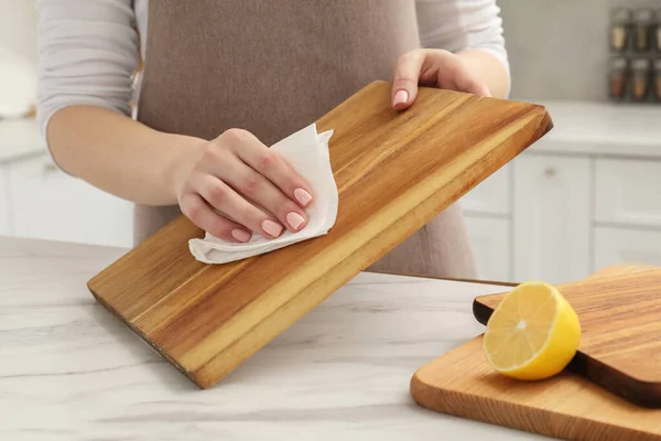 Woman wiping wooden cutting board with paper napkin at white table, closeup