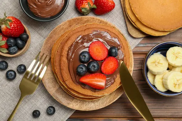 Tasty pancakes with chocolate paste, berries and banana served on wooden table, flat lay