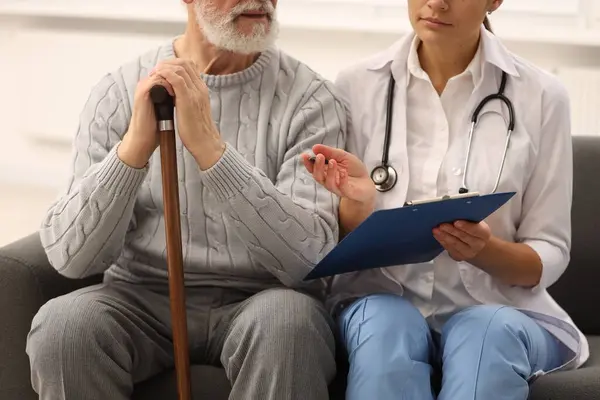 Nurse with clipboard assisting elderly patient on sofa indoors, closeup