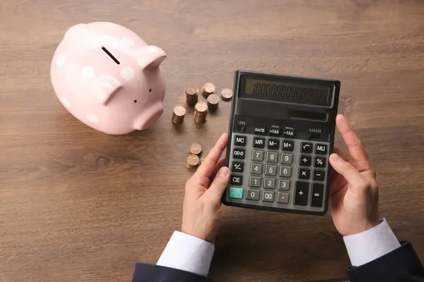 Budget planning. Businessman with piggy bank calculating at wooden table, closeup
