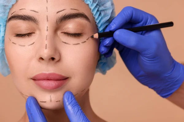 Doctor drawing marks on woman's face for cosmetic surgery operation against beige background, closeup