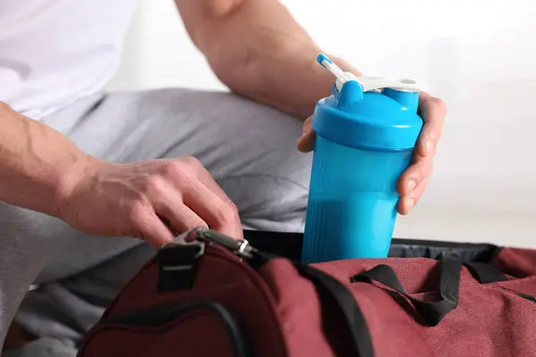 Young man putting shaker with protein into bag on blurred background, closeup
