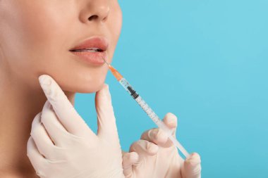 Doktor açık mavi arka planda genç bir kadına dudak enjeksiyonu yapıyor, yakın plan ve mesaj için boşluk. Estetik ameliyat