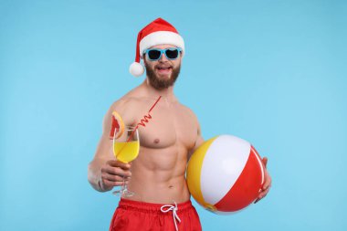 Noel Baba şapkalı kaslı çekici genç adam açık mavi arka planda top ve kokteyl tutuyor.