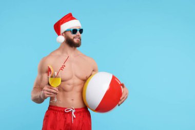 Noel Baba şapkalı çekici genç adam açık mavi arka planda top ve kokteyl tutuyor, mesaj için yer var.