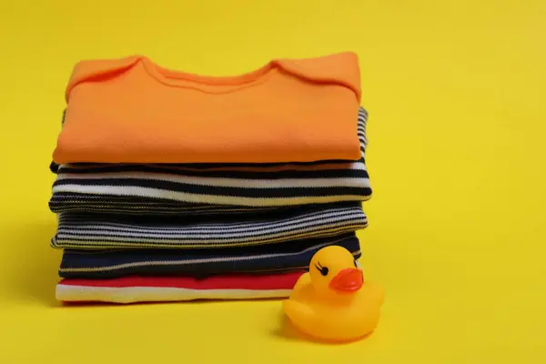 Stack of clean baby clothes and rubber duck on yellow background