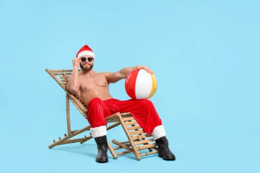 Noel Baba şapkalı kaslı genç adam, top, güneş gözlüğü ve açık mavi arka planda iskemle, mesaj için yer var.