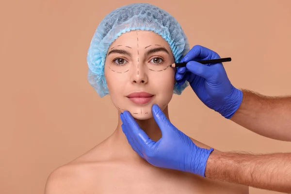 Doctor drawing marks on woman\'s face for cosmetic surgery operation against beige background
