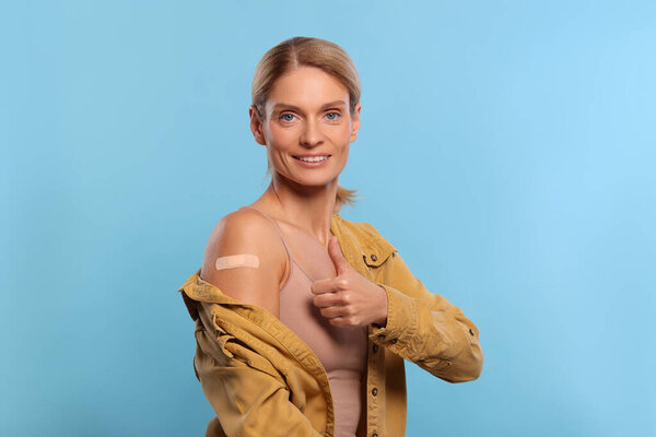 Smiling woman with adhesive bandage on arm after vaccination showing thumb up on light blue background