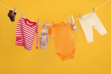 Farklı bebek kıyafetleri ve oyuncaklar turuncu arka planda çamaşır ipinde kuruyor.