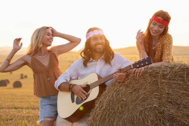 Güzel hippi kadınlar, arkadaşlarının tarlada gitar çalışını dinliyorlar.
