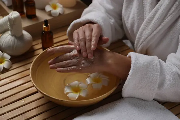 Woman applying scrub onto her hands in spa, closeup. Bowl of water and flowers on wooden table