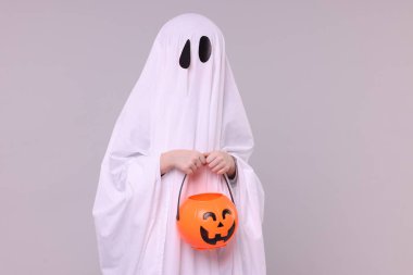 Beyaz hayalet kostümlü çocuk açık gri arka planda balkabağı kovası tutuyor. Cadılar Bayramı