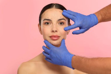 Doktor pembe arka planda estetik ameliyat yapmadan önce hastanın yüzünü kontrol ediyor.