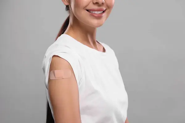 Woman with sticking plaster on arm after vaccination against light grey background, closeup