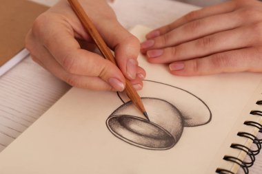 Masadaki çizim defterinde grafit kalemle içki bardağı çizen kadın, yakın plan.