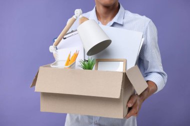 Mor arka planda kişisel ofis eşyalarıyla dolu kutusuyla işsiz genç adam, yakın plan.