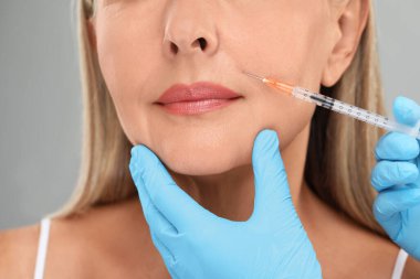 Doktor, açık gri arka planda yaşlı kadına yüz enjeksiyonu yapıyor. Yakın plan. Estetik ameliyat
