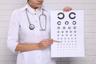 Oftalmolog, beyaz tuğla duvarın yakınındaki görme testi tablosuna işaret ediyor.