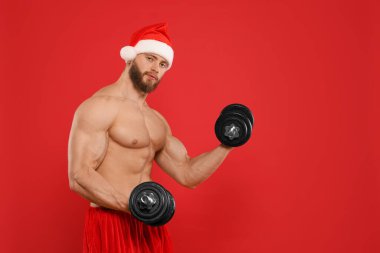Kırmızı arka planda halterleri olan, Noel Baba şapkalı kaslı genç adam, mesaj için yer var.