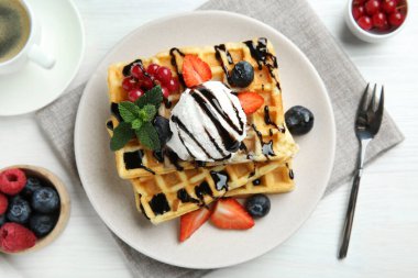 Beyaz ahşap masada servis edilen dondurma, böğürtlen ve çikolata soslu leziz Belçika waffleları.