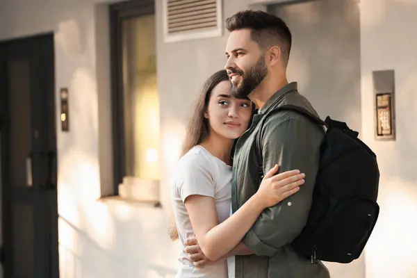 Long-distance relationship. Man with backpack hugging with his girlfriend near house entrance outdoors