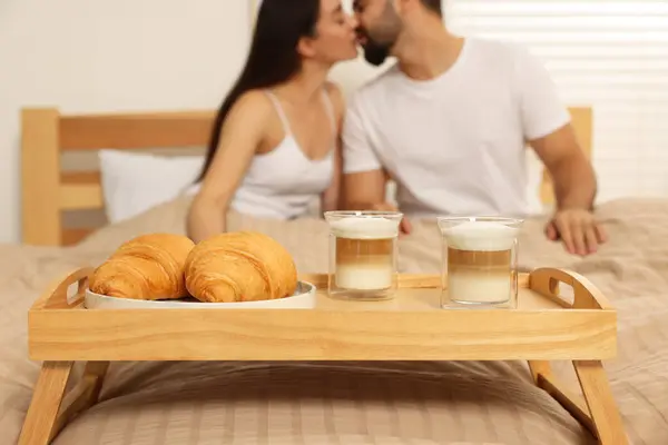 Happy couple kissing on bed at home, focus on wooden tray with breakfast