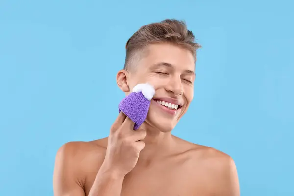 明るい青の背景にスポンジで顔を洗う幸せな若い男 — ストック写真
