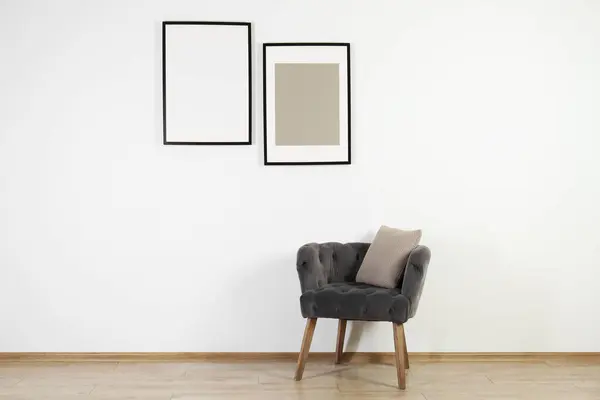 Comfortable armchair, cushion and frames in room with white wall. Interior design