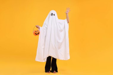 Beyaz hayalet kostümlü kadın sarı arka planda balkabağı kovası tutuyor. Cadılar Bayramı