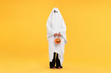 Beyaz hayalet kostümlü kadın sarı arka planda balkabağı kovası tutuyor. Cadılar Bayramı