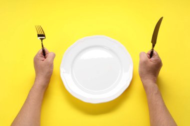 Çatallı, bıçaklı ve boş tabaklı adam sarı masada, üst tarafta.