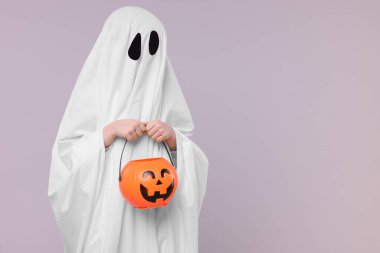 Beyaz hayalet kostümlü çocuk açık gri arka planda balkabağı kovası tutuyor, mesaj için yer var. Cadılar Bayramı